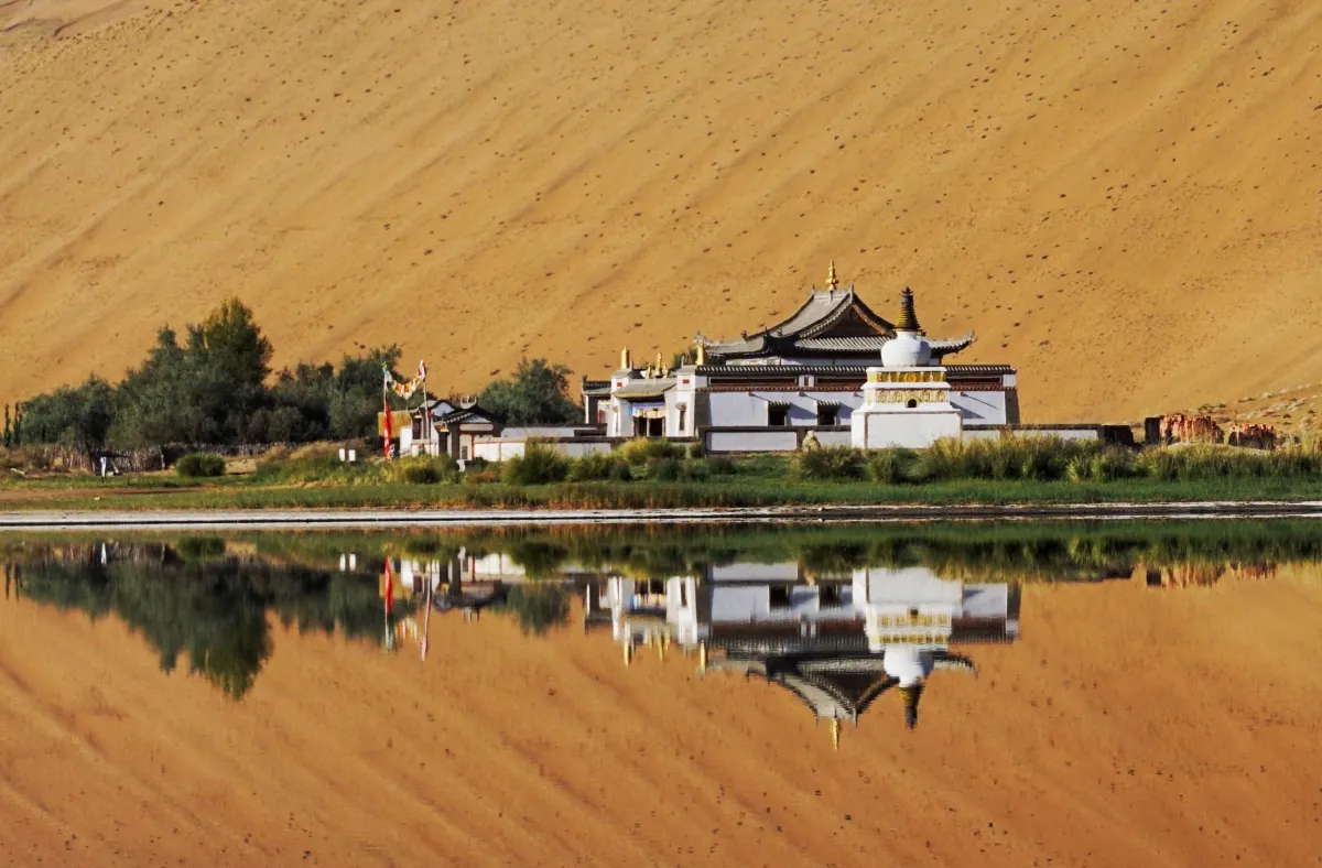 The historic white Tibetan monastery of Badain Jaran reflecting on a calm desert lake beneath towering sand peaks